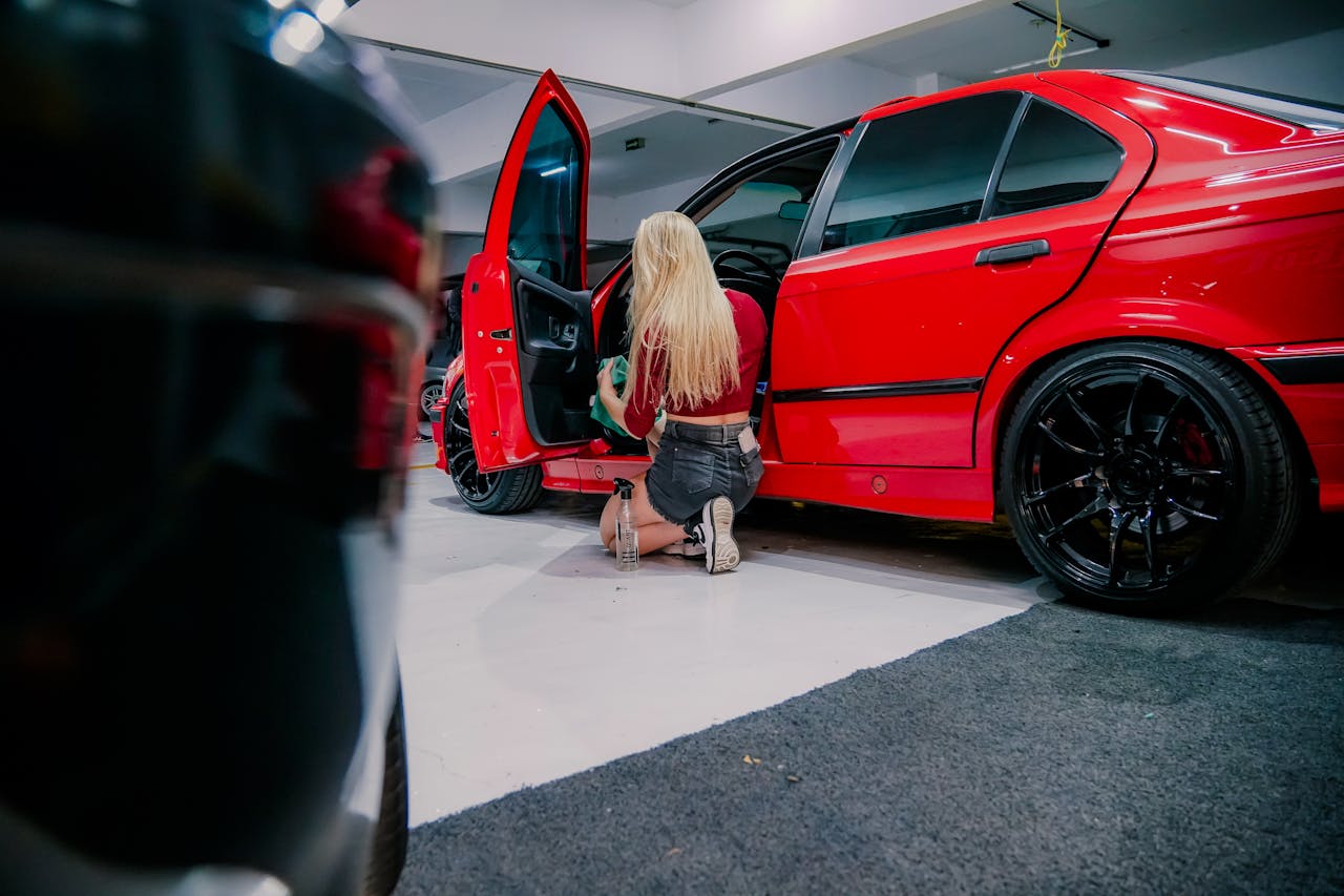 A woman kneels, cleaning the interior of a red BMW in a garage environment.